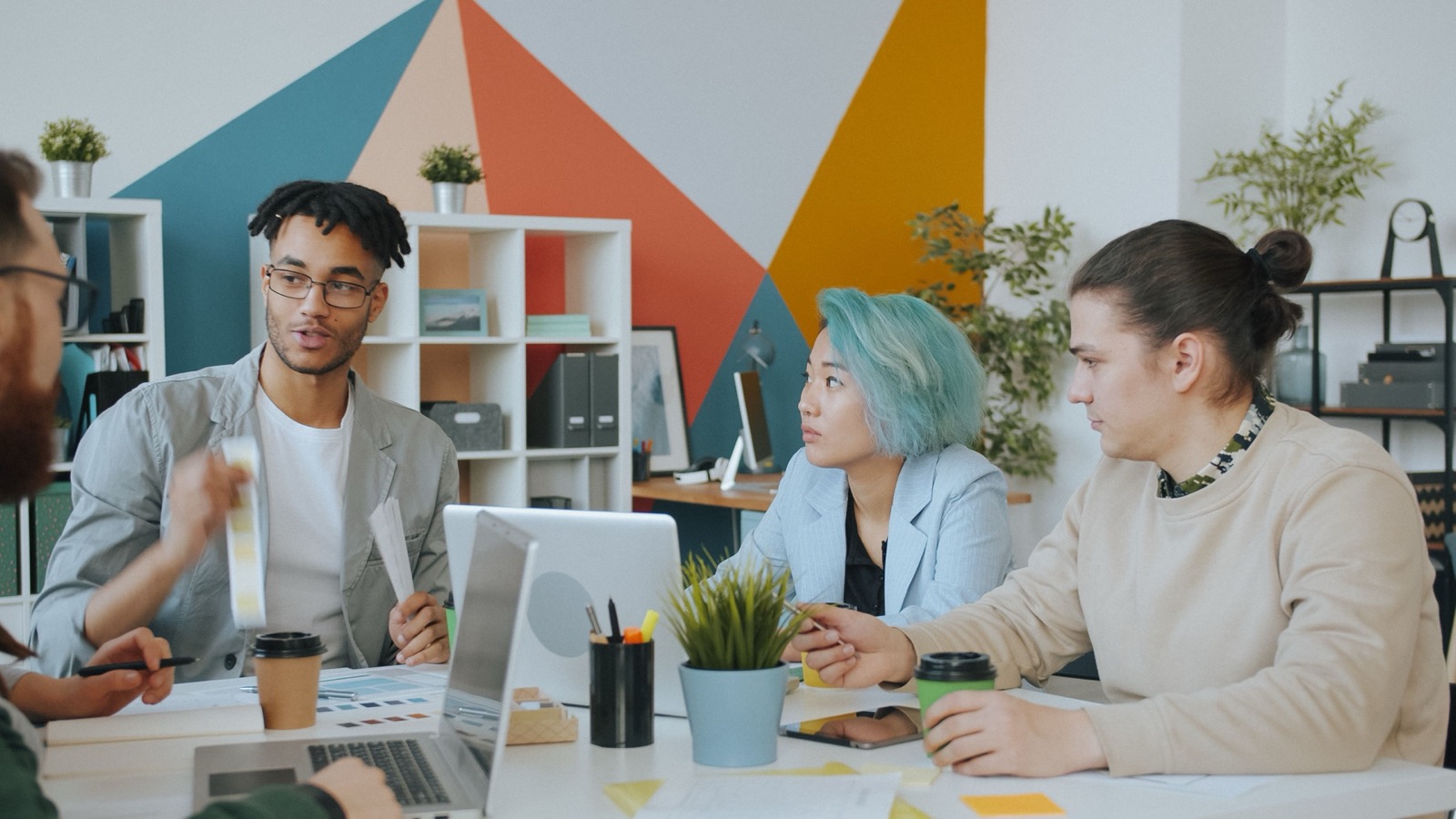 Four people around a table in a bright office, mid-conversation over laptops and coffee cups